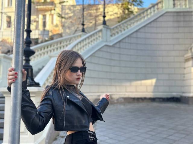 Bubochka with sunglasses, black leather crop jacket, and black skirt, holding a pole, showing her ass, with red nails, standing on a stone-paved area.