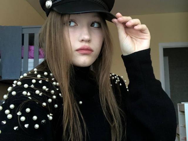 Bubochka mit langen braunen Haaren, blauen Augen und rosafarbenen Lippen, trägt eine schwarze Jacke und Mütze mit Nieten, berührt ihre Mütze, blickt zur Seite, in einem ungezwungenen Innenraum..
