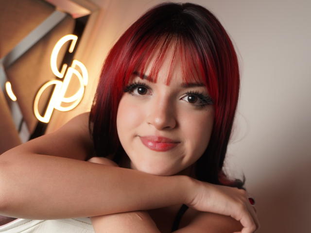IrisWithmore with red and black hair, smiling, has her arms crossed, showing her tits, and has long eyelashes, and red lipstick. Neon lights are in the background.