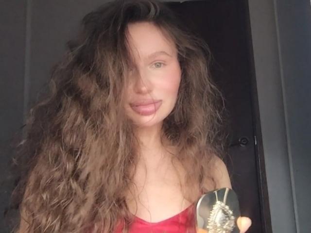 AnnPat with long, messy brown hair covering one eye, pink lips, and a red strapless top, holding a silver necklace, smiling softly.