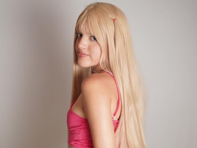 IrisWithmore heeft lang blond haar, een roze beha en een schattige glimlach. Ze kijkt over haar schouder, waarbij haar achterwerk zichtbaar is. Haar haar heeft een klein roze clipje..