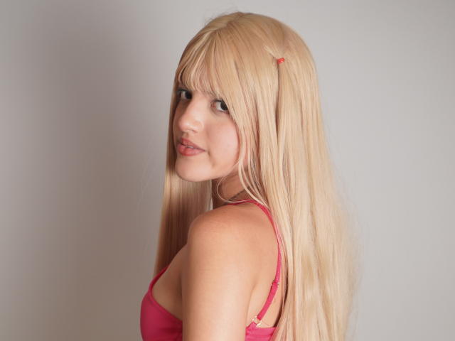 IrisWithmore heeft lang blond haar, een roze haarspeldje en een roze beha, en ze glimlacht naar de camera terwijl haar borsten lichtelijk zichtbaar zijn, terwijl ze over haar schouder kijkt..