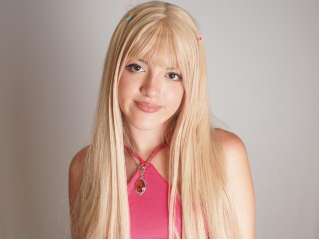 IrisWithmore has long blonde hair with bangs, brown eyes, and a pink halter top with a red necklace. She's smiling softly with a cute, youthful look.