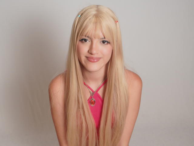 IrisWithmore has long blonde hair, pink halter top, and a cute smile. She's wearing a pink necklace with a heart pendant, looking forward. Her tits are small.