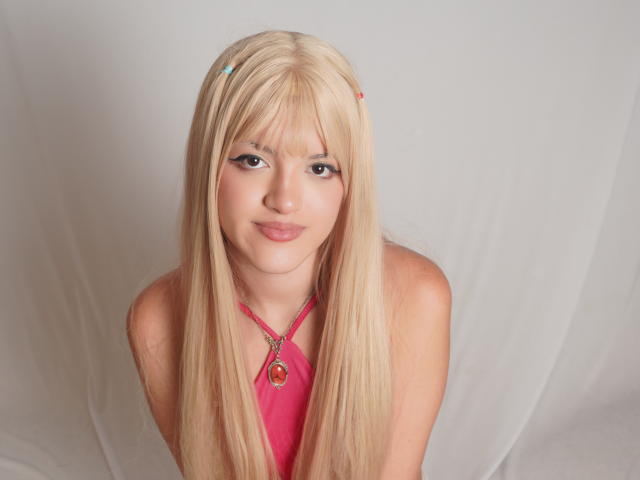 IrisWithmore has long blonde hair, a pink halter top, and a necklace with a red pendant. She's smiling with a cute face and small tits. 

