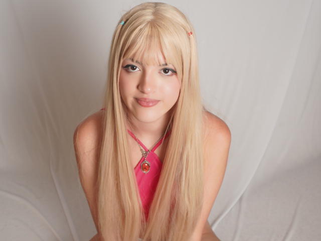 IrisWithmore has long blonde hair, blue hairpins, and a pink halter top, smiling with her tits barely visible, sitting against a white background.