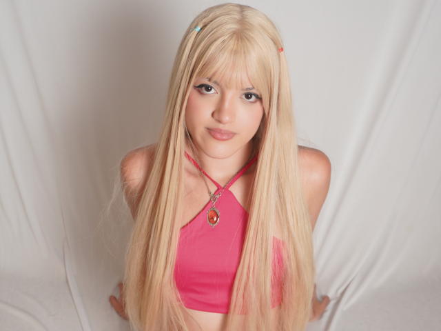 IrisWithmore has long blonde hair, pink halter top, and a necklace with a red gem. She's smiling with a cute face, looking up at the camera.