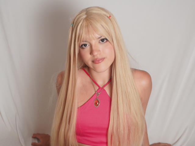 IrisWithmore has long blonde hair, a pink halter top, and a red necklace with a gem. She's smiling at the camera with a cute, sexy look.