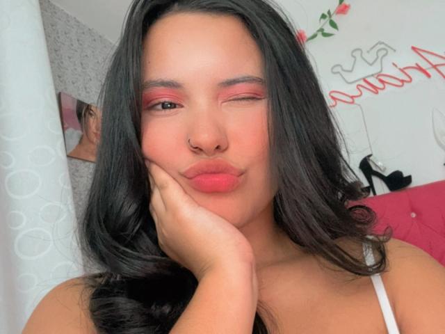 ArianaSmith69 with wavy black hair, pink eyeshadow, nose ring, puckered lips, hand on cheek, white tank top, and a cute, playful expression. Her pussy is not visible.