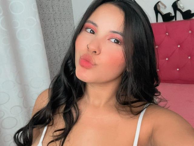 ArianaSmith69 with long black hair, pink eyeshadow, nose ring, pouty pink lips, white spaghetti strap top, showing her tits, pink tufted headboard, black high heels in background.