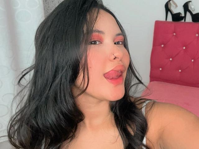 ArianaSmith69 with black hair, nose ring, and red eyeshadow, tongue out, wearing a white tank top, sitting on a pink bed with black high heels on the headboard.