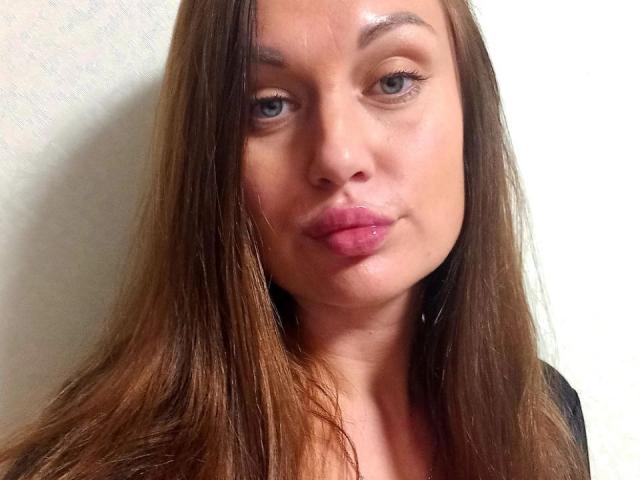 SuzieJam, con cabello castaño largo, labios rosados y ligeramente curvados, y una leve sonrisa, mirando directamente a la cámara con ojos azules y un maquillaje sutil..