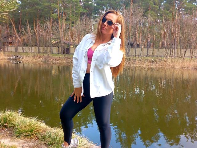 SuzieJam, con cabello rubio largo, usando gafas de sol, una camisa blanca abotonada, un sujetador rosa y leggings negros, de pie junto a un estanque con una mano en la cabeza..
