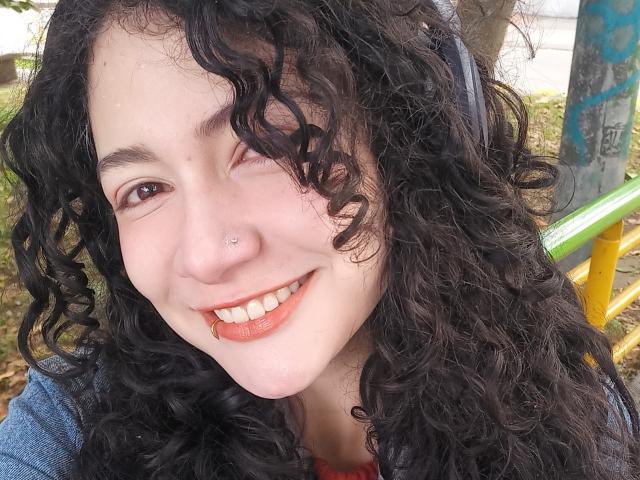 AmeliaTorres has curly black hair, a nose piercing, and a smile with orange lipstick, showing her white teeth. She's wearing a blue jacket.