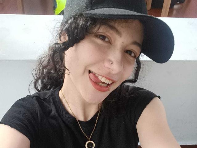 AmeliaTorres with curly black hair, black cap, black shirt, gold necklace, and nose ring, sticking out her tongue. She has a small nose piercing and light skin.