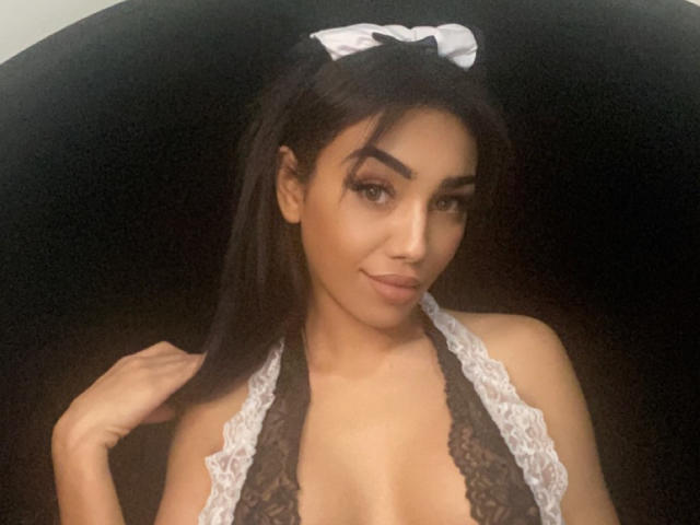 BellaRani with dark hair in a ponytail, lace halter top, and white headband, showing her tits, smiling, and touching her hair. Soft lighting, confident expression.