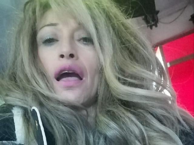 BigBoobsy69 with long, messy blonde hair, pink eyeshadow, and open mouth, looking slightly dazed or shocked, wearing a black and white hoodie.