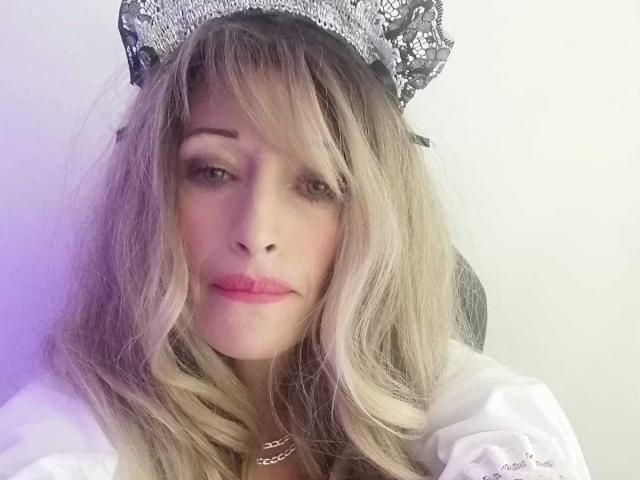 BigBoobsy69 with blonde wavy hair, wearing a silver sequined headpiece, pink lipstick, and a white shirt, looking directly at the camera with a slightly serious expression.