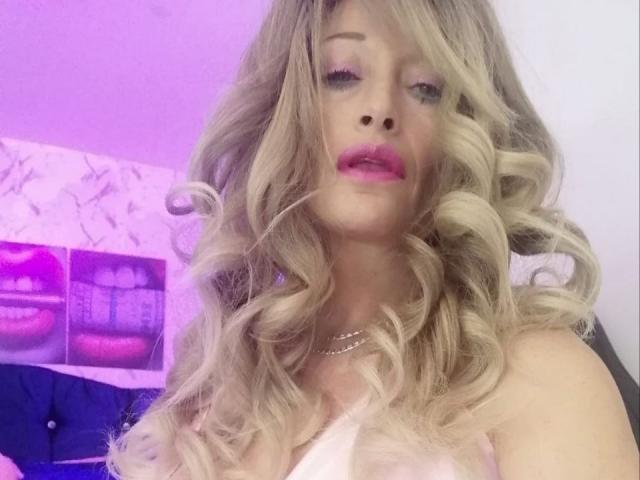 Blonde BigBoobsy69 with curly hair, pink lipstick, and a necklace, gazing forward with a slightly open mouth. Purple and pink lighting in the background.