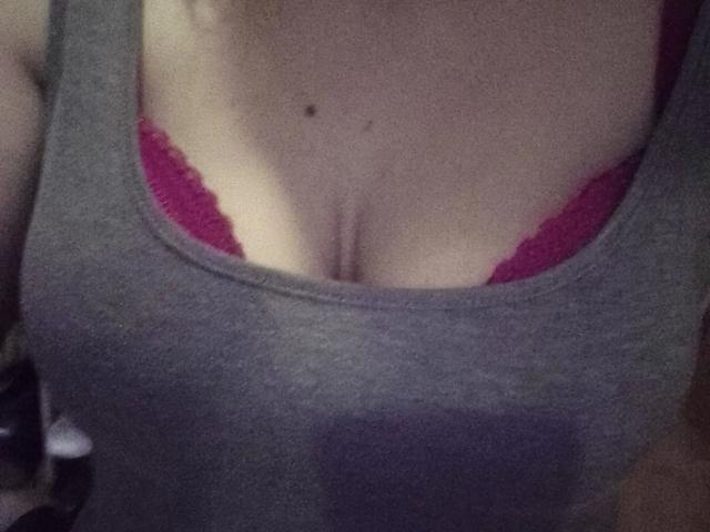 Sexybienchen93-hot's tits, covered in a gray tank top, with red lace bra visible at the top. Small mole above cleavage. Soft lighting, subtle shadow.