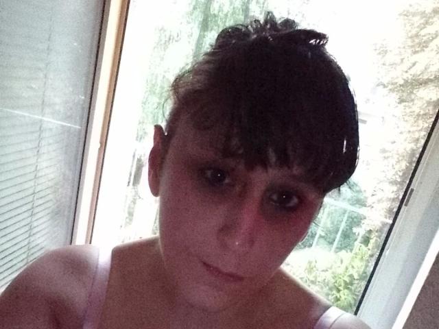 Sexybienchen93-hot with short brown hair, dark eye makeup, and a pink strap, looking down with a slightly confused expression, standing near a window.