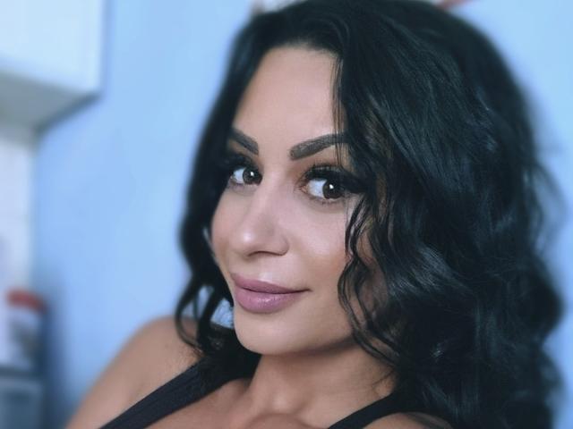 SweetyFinger has wavy black hair, brown eyes, and a subtle smile. She's wearing a black top with visible shoulders, looking cute and attractive. Her ass is not visible.