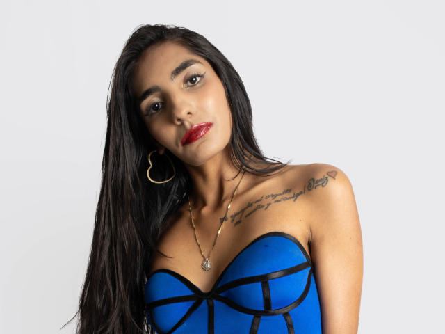 JuicyRoseX with long black hair, blue strapless top, red lipstick, gold hoop earrings, necklace, tattoo on right shoulder, looking at camera, slightly tilted head, white background.