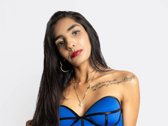 JuicyRoseX with long black hair, red lipstick, hoop earrings, blue strapless top, tattoo on right shoulder, looking at camera, necklace with pendant, shiny skin.