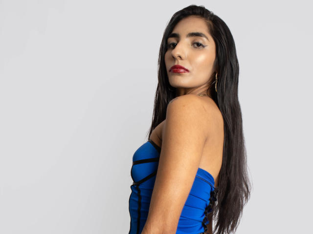 JuicyRoseX with long black hair, blue strapless dress, red lipstick, and gold earrings, looking back over her shoulder, showing her tits.