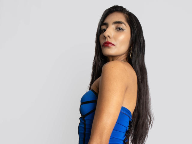 JuicyRoseX with long black hair, red lipstick, and blue strapless dress, looking over shoulder, showing bare shoulders and small, firm tits. She has tan skin and gold earrings.