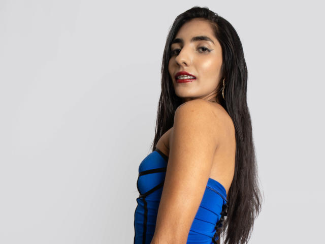 JuicyRoseX with long black hair, blue strapless dress, and red lipstick, looking over shoulder, showing her tits, and a slight smile, against a plain white background.