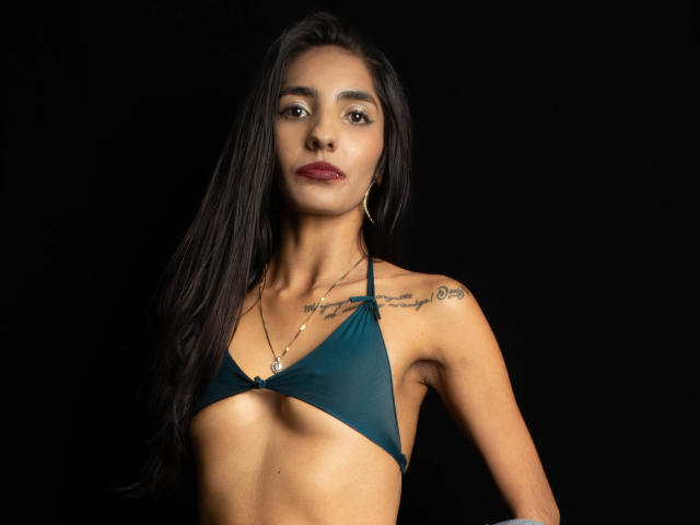 JuicyRoseX with dark hair, wearing a teal bikini top, showing small tits, tattoos on her shoulder, gold hoop earrings, and a necklace, against a black background.
