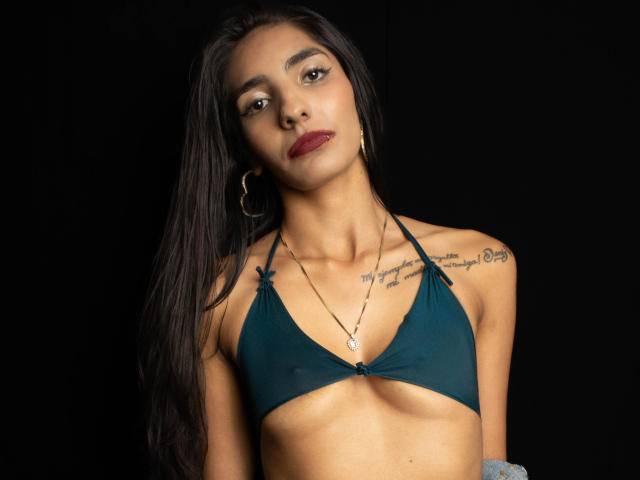 JuicyRoseX with long black hair, dark green bikini top, small tits, tattoos on chest, gold hoop earrings, red lipstick, necklace, black background.