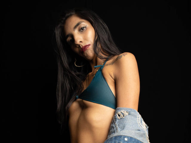 JuicyRoseX with dark hair, wearing a teal bikini top, gold hoop earrings, and a blue ripped jacket, showing tattoos on her shoulder, against a black background.