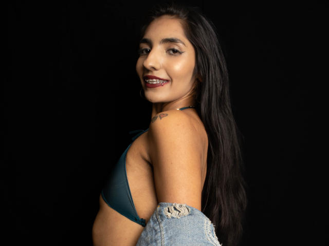 JuicyRoseX with long black hair, dark lipstick, wearing a teal bra, denim jacket off, smiling, tattoo on right shoulder, black background, brown skin, small breasts, side profile.