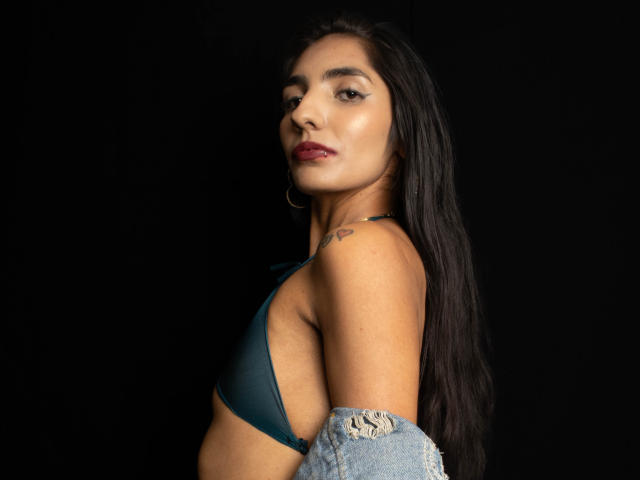 JuicyRoseX with long black hair, dark red lipstick, wearing a green bra, holding a torn blue shirt, showing a small heart tattoo on her right shoulder.