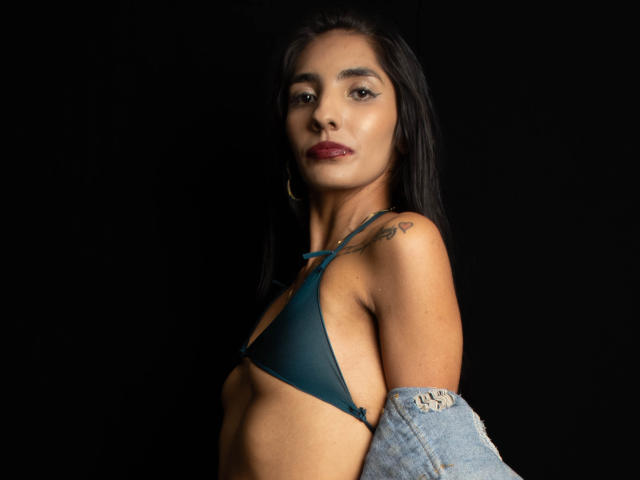 JuicyRoseX with dark hair, medium tits in a teal bikini, tattoo on right shoulder, wearing a denim jacket, black background, red lipstick, and hoop earrings.