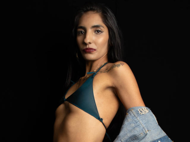 JuicyRoseX with dark hair, wearing a teal bra, denim jacket off, showing small tits and a tattoo on her right shoulder, standing against a black background.