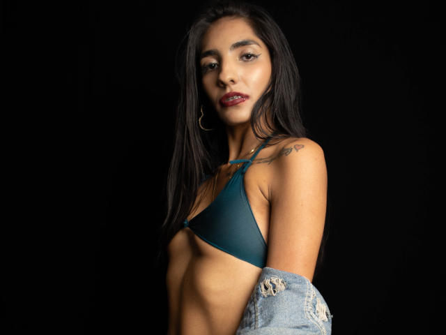 JuicyRoseX with long black hair, dark eyes, and red lipstick, wearing a teal bikini top, showing small tits, tattoos on her shoulder, and a blue jacket off her arms.