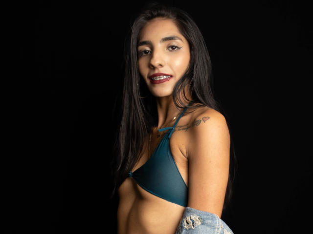 JuicyRoseX with long black hair, wearing a green bra, showing small tits, tattoo on right shoulder, light brown skin, dark red lipstick, black background, and denim jacket.
