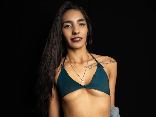 JuicyRoseX with long black hair, brown skin, small tits in a dark green bikini top, tattoos on shoulders, wearing gold earrings and necklace, smiling against a black background.