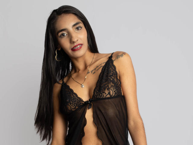 JuicyRoseX with long black hair, wearing a sheer black lace top, showing small tits, tattoos on shoulders, red lipstick, gold hoop earrings, and a necklace against a gray background.