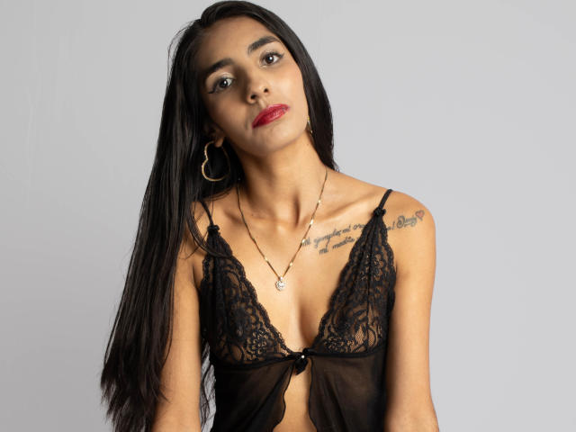 JuicyRoseX with long black hair, wearing a sheer black lace bralette, showing small tits and tattoos on her right shoulder, red lipstick, gold hoop earrings, and a necklace.