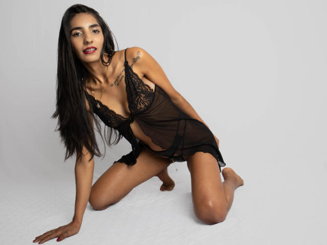 JuicyRoseX is kneeling on the floor, wearing a black lace lingerie dress, exposing her small tits and black panties, with long black hair and a tattoo on her right shoulder.
