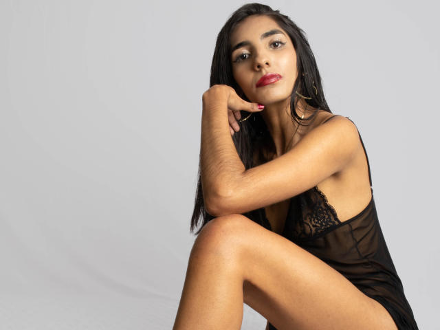 JuicyRoseX with dark hair, red lipstick, and black lace lingerie, sitting with one leg up, hand on chin, looking at camera, wearing gold hoop earrings, showing her tits.