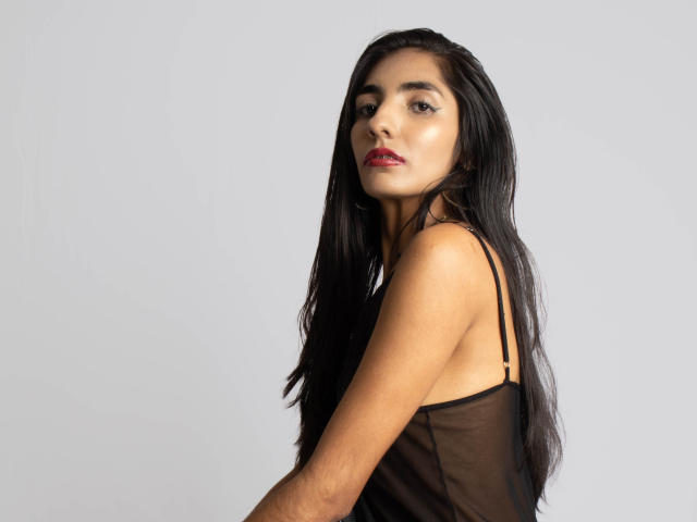 JuicyRoseX has long black hair, wearing a sheer black top, showing her ass and looking over her shoulder with red lips and dark eyeliner.