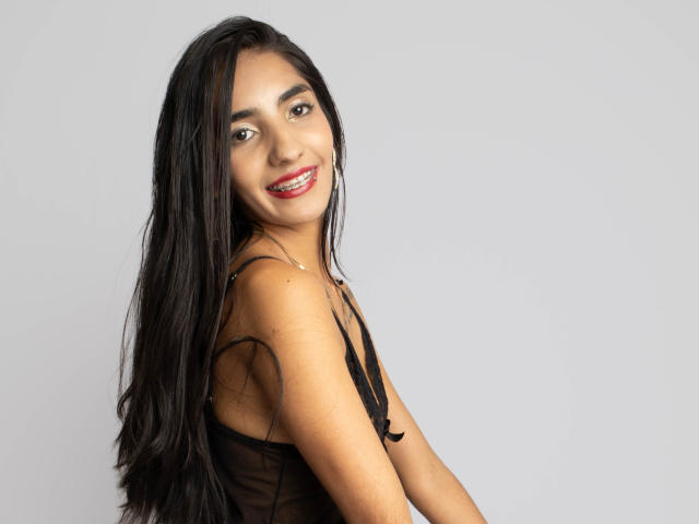 JuicyRoseX with long black hair, smiling, wearing a black sheer top, showing her small tits, standing against a plain grey background, wearing red lipstick and hoop earrings.