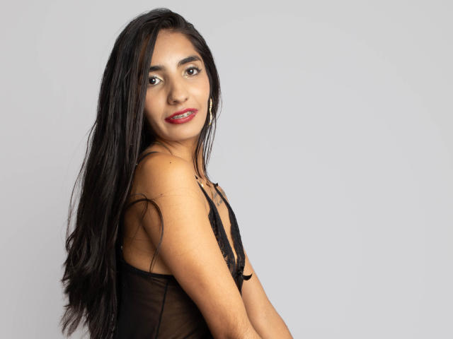JuicyRoseX with long black hair, wearing a sheer black top, showing small tits, smiling with red lipstick, standing against a plain grey background.
