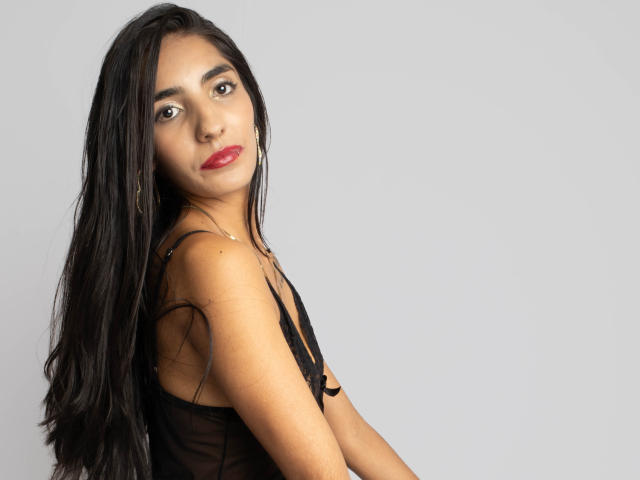 JuicyRoseX has long black hair, red lipstick, and a serious expression. She's wearing a black sheer top, showing her small, perky tits. She's facing sideways, looking at the camera.