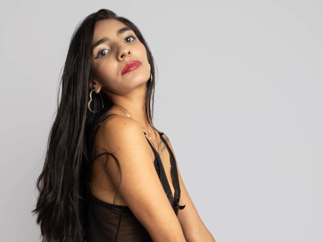 JuicyRoseX with long black hair, red lips, hoop earrings, and black sheer top, looking up with a slight tilt, showing her bare shoulders and small, perky tits.